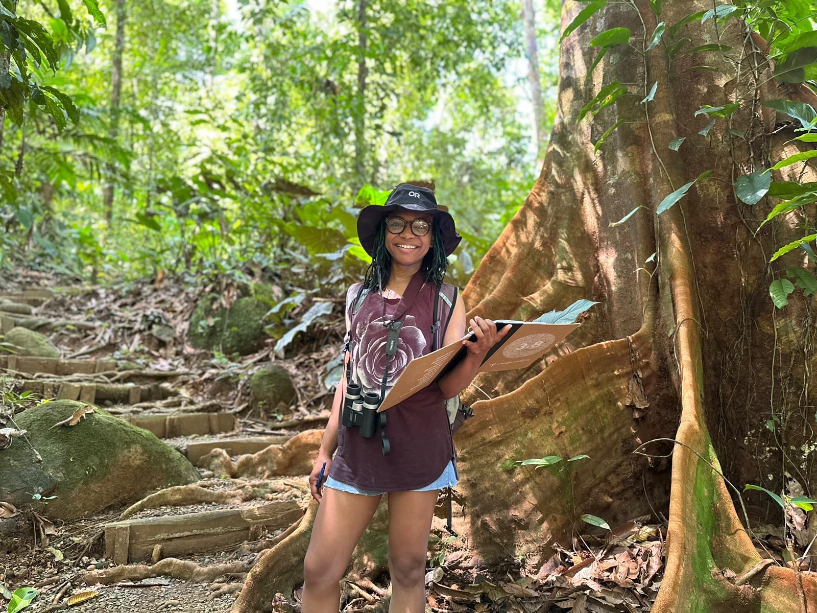 Genell standing beside the buttress roots of a giant rainforest tree, holding a field notebook.